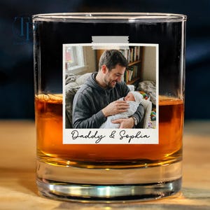 May include: A rocks glass filled with amber liquid, featuring a photo of a man holding a baby, taped to the glass. The photo has the text "Daddy & Sophia" at the bottom. The glass sits on a wooden surface.