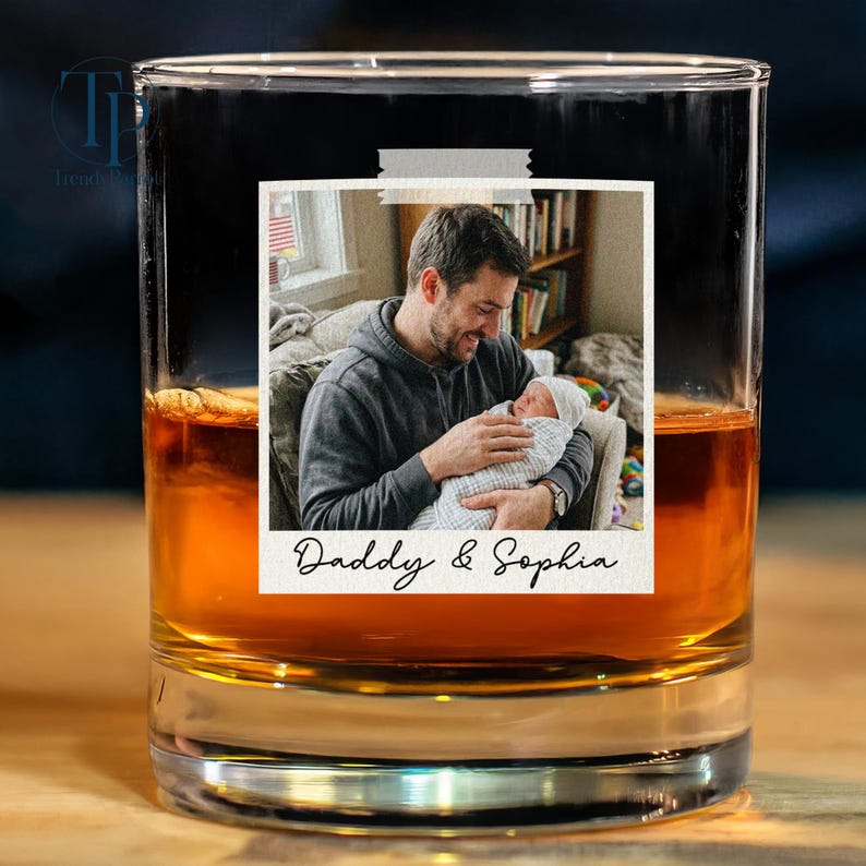 May include: A rocks glass filled with amber liquid, featuring a photo of a man holding a baby, taped to the glass. The photo has the text "Daddy & Sophia" at the bottom. The glass sits on a wooden surface.