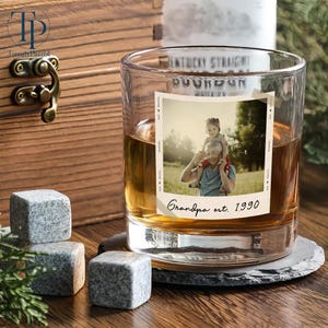 May include: A rocks glass with a photo of a person and child, the text "Grandpa est. 1990", and whiskey. Beside the glass are three gray granite whiskey stones. A wooden box is in the background.