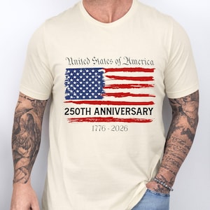 May include: Cream-colored t-shirt featuring a distressed American flag design in red, white, and blue. The text "United States of America" is above the flag, with "250TH ANNIVERSARY 1776-2026" below. The shirt is short-sleeved.
