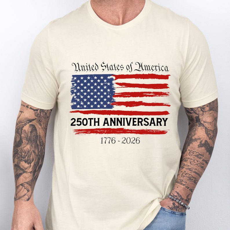 May include: Cream-colored t-shirt featuring a distressed American flag design in red, white, and blue. The text "United States of America" is above the flag, with "250TH ANNIVERSARY 1776-2026" below. The shirt is short-sleeved.