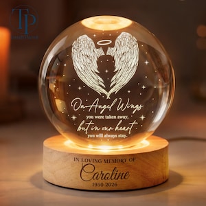 May include: A lit crystal sphere featuring angel wings and the text "On Angel Wings" on a wooden base. The base is engraved with "In Loving Memory of Caroline 1950-2026". The sphere emits a warm, soft light.