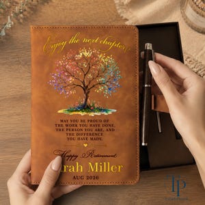 May include: A brown leather-look journal featuring a colorful tree illustration, the text "Enjoy the next chapter," a retirement quote, the name "Sarah Miller," and "AUG 2026." A dark brown metal pen is positioned in the adjacent open gift box.