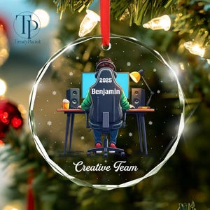 May include: A clear, faceted glass Christmas ornament with a red ribbon. The ornament features a gamer design with a person sitting at a desk with a computer, speakers, and the text "2025 Benjamin" and "Creative Team".