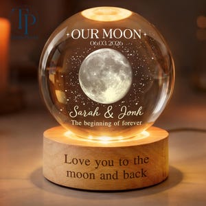 May include: A clear glass sphere lamp featuring an internal 3D moon and star design. The text reads OUR MOON, 06.03.2026, Sarah & Jonh, and The beginning of forever. It sits on a light wood base engraved with Love you to the moon and back.