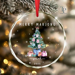 May include: A clear, round Christmas ornament with a red ribbon. The ornament features a watercolor Christmas tree decorated with Mahjong tiles, gifts, and the text "MERRY MAHJONG" and "Sarah 2025".