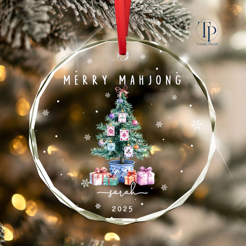 May include: A clear, round Christmas ornament with a red ribbon. The ornament features a watercolor Christmas tree decorated with Mahjong tiles, gifts, and the text "MERRY MAHJONG" and "Sarah 2025".