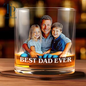 May include: A clear whiskey glass with a photo of a man and two children inside. The words "BEST DAD EVER" are printed in white on the glass. The glass sits on a wooden coaster.