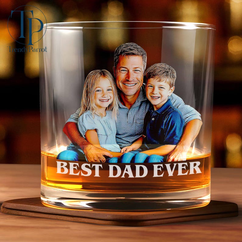 May include: A clear whiskey glass with a photo of a man and two children inside. The words "BEST DAD EVER" are printed in white on the glass. The glass sits on a wooden coaster.