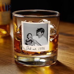 May include: A rocks glass filled with amber liquid, ice cubes, and a vintage photo. The photo shows a man holding a baby, with the text "Dad est. 1996" below. The glass sits on a wooden surface.