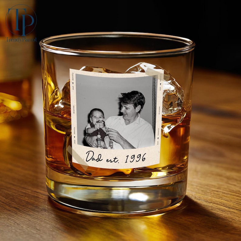 May include: A rocks glass filled with amber liquid, ice cubes, and a vintage photo. The photo shows a man holding a baby, with the text "Dad est. 1996" below. The glass sits on a wooden surface.