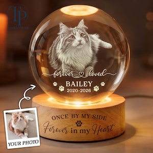 May include: A clear glass sphere engraved with a cat image, the text "forever loved", "BAILEY", and "2020-2026" with paw prints. It sits on a round wooden base engraved with "ONCE BY MY SIDE Forever in my Heart" and a paw print icon.