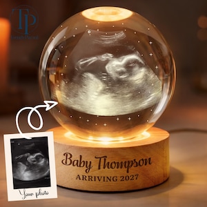 May include: A spherical glass orb featuring an internal laser-etched ultrasound image of a fetus, illuminated by a light-up wooden base. The base is engraved with the text Baby Thompson ARRIVING 2027. A small square photo print shows the original ultrasound.