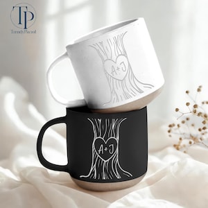 May include: Two ceramic mugs, one white and one black, stacked. Each mug features a tree design with a heart and the initials "A+J". The mugs have a neutral-colored base and a curved handle. The logo "TrendyParrot" is in the top left corner.