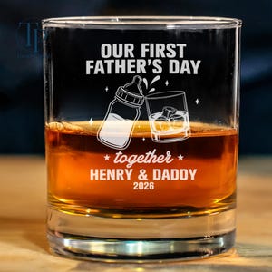 May include: A clear glass tumbler filled with amber liquid, possibly whiskey, with ice cubes. The glass is etched with the words "OUR FIRST FATHER'S DAY" and the names "HENRY & DADDY" with the year 2026. A baby bottle and a glass of whiskey are illustrated.