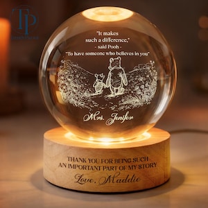 May include: A crystal sphere illuminated from within, resting on a wooden base. The sphere displays an etched scene of Winnie the Pooh and Piglet, accompanied by quotes. The base is engraved with a personalized message, creating a warm glow.