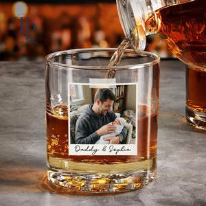May include: A clear rocks glass with a personalized photo of a man holding a baby, with the text "Daddy & Sophia." Amber liquid is being poured into the glass from a decanter. The glass is partially filled with amber liquid.