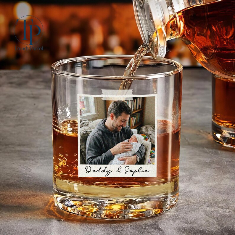 May include: A clear rocks glass with a personalized photo of a man holding a baby, with the text "Daddy & Sophia." Amber liquid is being poured into the glass from a decanter. The glass is partially filled with amber liquid.