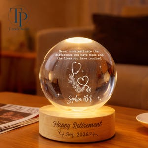May include: A clear glass sphere lamp on a round wooden base. The sphere features an etched stethoscope, floral design, and the text: Never underestimate the difference you have made and the lives you have touched. Sophia, RN. Base reads: Happy Retirement Sep 2026.