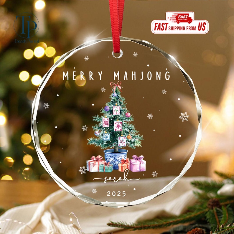 May include: A clear, round Christmas ornament with a red ribbon. The ornament features a Christmas tree decorated with Mahjong tiles, gifts, and the text "MERRY MAHJONG" and "Sarah 2025".
