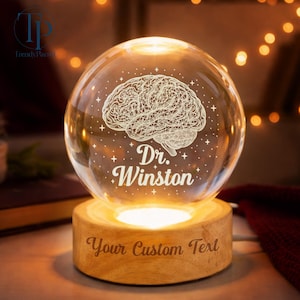 May include: A lit glass sphere featuring a brain design and the name "Dr. Winston". The sphere rests on a wooden base with the words "Your Custom Text". The internal illumination casts a warm glow, creating a decorative and personalized item.