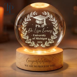 May include: A glowing crystal sphere with a graduation cap design, the text "Ph.D Dr. Lisa Evans University of Michigan 2026" is etched inside. The sphere sits on a wooden base with the words "Congratulations! WE ARE SO PROUD OF YOU!"