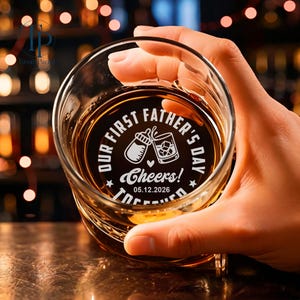 May include: A clear whiskey glass with a personalized design. The design includes the text "OUR FIRST FATHER'S DAY" and "Cheers!" along with a baby bottle and a whiskey glass. The date "05.12.2026" is also included. The glass is filled with amber liquid.