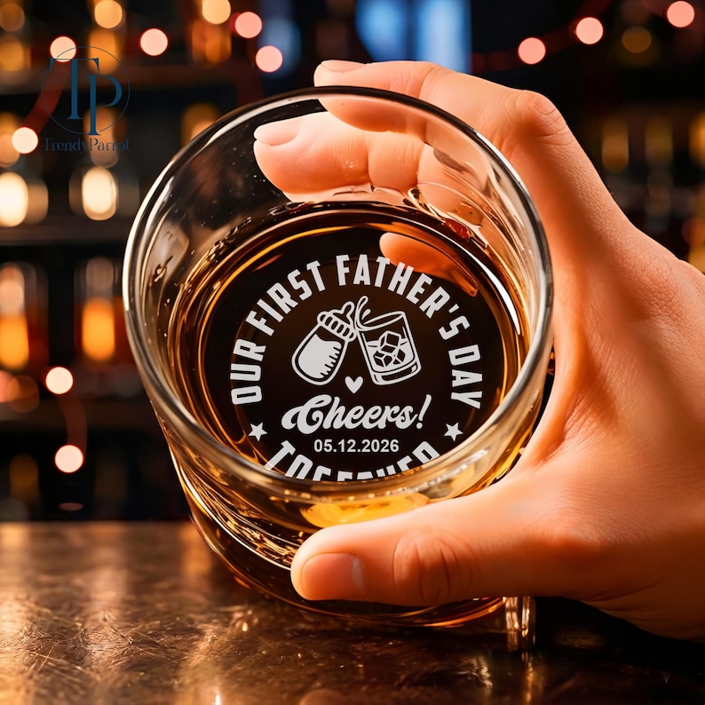 May include: A clear whiskey glass with a personalized design. The design includes the text "OUR FIRST FATHER'S DAY" and "Cheers!" along with a baby bottle and a whiskey glass. The date "05.12.2026" is also included. The glass is filled with amber liquid.