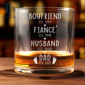 May include: A clear glass tumbler with the words "Boyfriend Est. 1993", "Fiance Est. 1998", "Husband Est. 2000", and "Dad Est. 2015" etched on the side. The glass is filled with amber liquid.