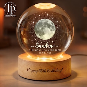 May include: A spherical glass night light featuring an engraved moon and star pattern, the name Sandra, and the text THE NIGHT YOU WERE BORN 06.03.1966. The glass sphere rests on a cylindrical wooden base engraved with Happy 60th Birthday.