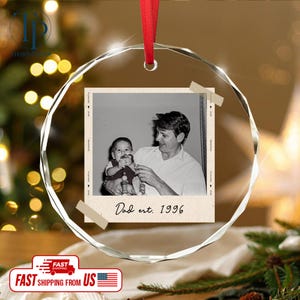 May include: A clear, faceted glass ornament with a red ribbon. Inside is a black and white photo of a man holding a baby. The photo is labeled "Dad est. 1996".