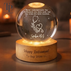 May include: A clear glass sphere night light on a round wooden base. The sphere is engraved with a stethoscope, flowers, stars, and the text: Never underestimate the difference you have made and the lives you have touched, Sophia, RN. The base reads: Happy Retirement Sep 2026.