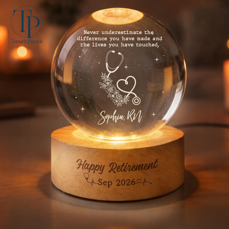 May include: A clear glass sphere night light on a round wooden base. The sphere is engraved with a stethoscope, flowers, stars, and the text: Never underestimate the difference you have made and the lives you have touched, Sophia, RN. The base reads: Happy Retirement Sep 2026.