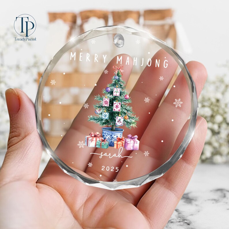 May include: A clear, round ornament with the words "MERRY MAHJONG" and "Sarah 2025" printed on it. The ornament features a Christmas tree decorated with Mahjong tiles, ornaments, and gifts. The ornament is held in a hand.