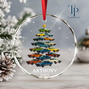 May include: A clear, round Christmas ornament with a red ribbon. The ornament features a tree made of colorful classic cars, topped with a gold star. The name "ANTHONY" and the year "2025" are printed at the bottom.