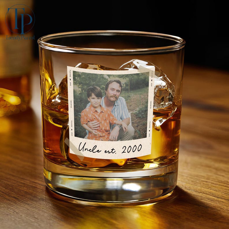 May include: A rocks glass filled with amber liquid and ice cubes, featuring a photo with a man and a child. The photo has the text "Uncle est. 2000" at the bottom. The glass sits on a wooden surface.