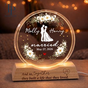 May include: A circular acrylic plaque with LED illumination set in a wooden base. The plaque features white floral illustrations, a silhouette of a couple, and the text: Molly, Henry, married, May 27, 2026. The base is engraved: And so, together they built a life that they loved.