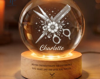 May include: A clear glass sphere lamp resting on a cylindrical wooden base. The sphere features an etched design of scissors, a comb, and a flower with the name Charlotte. The base is engraved with the text: NEVER UNDERESTIMATE THE DIFFERENCE YOU MAKE AND THE LIVES YOU TOUCH.