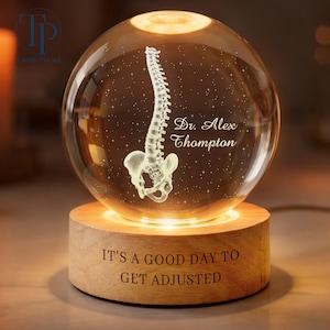 May include: A clear glass sphere resting on a cylindrical wooden base. The sphere features an internal laser-etched human spine and pelvis with the text Dr. Alex Thompton. The wooden base is engraved with the text IT'S A GOOD DAY TO GET ADJUSTED.