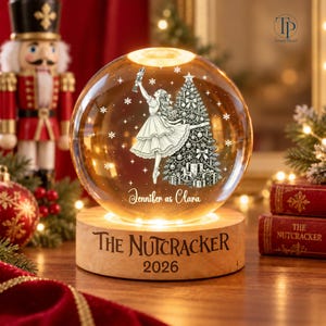 May include: A spherical glass ornament with an internal light, featuring a printed illustration of a ballerina dancing next to a decorated Christmas tree. The text reads Jennifer as Clara, The Nutcracker, and 2026 on the wooden base.