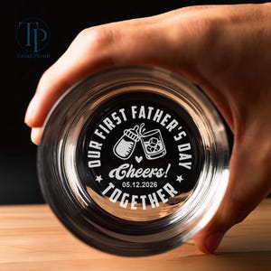 May include: A clear glass with the text "OUR FIRST FATHER'S DAY" and "Cheers!" encircling a baby bottle and a cocktail glass. The date "05.12.2026" and the word "TOGETHER" are also included. The glass is held by a hand.