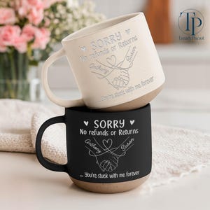 May include: Two ceramic mugs, one cream and one black, stacked. The cream mug has a white handle and the black mug has a black handle. Both mugs have the text "SORRY No refunds or Returns" with names and a heart graphic. The bottom of each mug is a speckled brown.