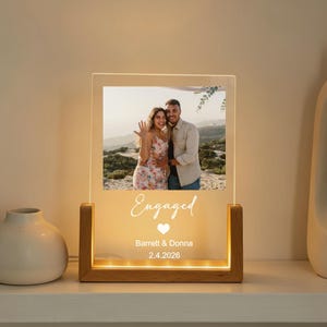 Könnte beinhalten: Ein beleuchteter Acryl-Fotorahmen mit Holzsockel, der ein Verlobungsfoto eines Paares zeigt. Das Wort "Engaged" steht auf dem Rahmen, zusammen mit den Namen des Paares und dem Datum 2.4.2026. Der Rahmen wird von warmweißen Lichtern beleuchtet.