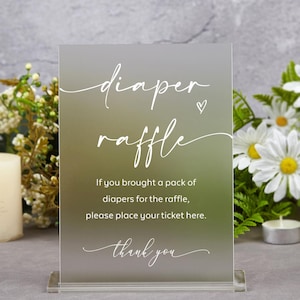 May include: A clear acrylic sign with white text that reads "diaper raffle". The sign also includes the text "If you brought a pack of diapers for the raffle, please place your ticket here. Thank you."