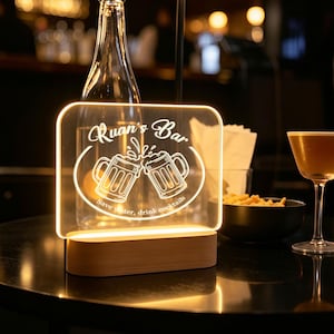 Könnte beinhalten: Ein rechteckiges, beleuchtetes Acrylschild mit Holzsockel, das den Text Ruan's Bar und eine Grafik von zwei anstoßenden Bierkrügen mit dem Text Save water, drink cocktails zeigt. Das Schild strahlt ein warmes Licht aus und hebt das Design hervor.