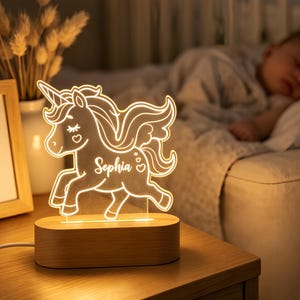 May include: A clear acrylic night light shaped like a winged unicorn with the name Sophia engraved on its body. The unicorn features a flowing mane, a horn, and a small heart. It is mounted on a smooth, oval-shaped wooden base with a power cord.