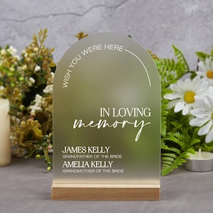Puede incluir: Un letrero de acrílico transparente con la parte superior redondeada y una base de madera. El letrero dice "Wish you were here" en un círculo en la parte superior y "In loving memory" con los nombres "James Kelly, Grandfather of the Bride" y "Amelia Kelly, Grandmother of the Bride" debajo.