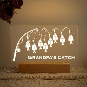 May include: A personalised night light with a fishing theme. The clear acrylic panel features a fishing rod design with fish, each labelled with a name. The base is made of wood, and the text "GRANDPA'S CATCH" is displayed.