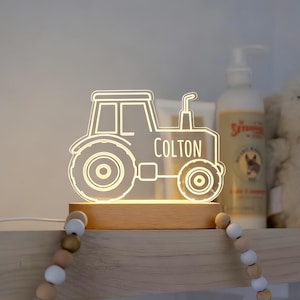 May include: A tractor-shaped night light with the name "COLTON" illuminated in white. The light sits on a wooden base and is plugged into an electrical outlet. The light is on a shelf with a decorative beaded garland.