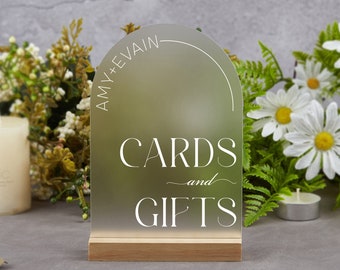May include: A clear acrylic sign with a rounded top and a wooden base. The sign says "Amy + Evian" in a circle at the top and "Cards and Gifts" in the center.